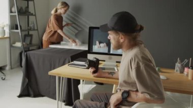 Beyzbol şapkalı, sıradan tişörtlü, kırmızı sakallı beyaz bir adamın orta boy portresi. Bilgisayar monitörünün önünde oturmuş, elinde ürün örneği fotoğraflarıyla kameraya bakıyor.