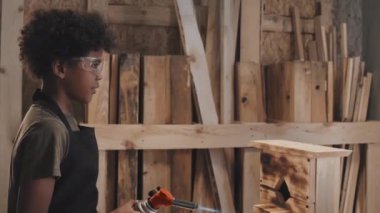 Babası marangozluk atölyesinde onu desteklerken Afrika kökenli Amerikalı çocuk ahşap kuş evinin yüzeyini yakmak için özel bir alet kullanıyor.
