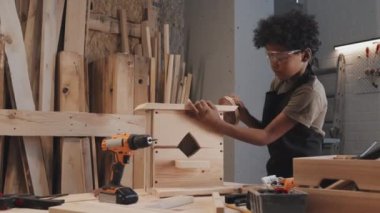 Afro-Amerikan çocuk marangozluk atölyesinde çalışırken kuş yuvasının yüzeyini zımparalıyor.