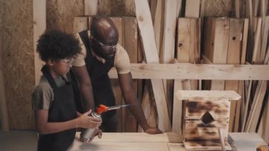 Afrikalı Amerikalı baba, oğullarına marangozluk atölyesinde çalışırken kuş evini yakmayı öğretiyor.