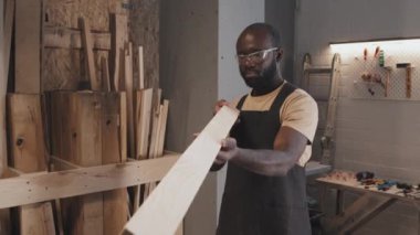 Afrikalı Amerikan marangoz elinde büyük bir kalas tutuyor ve marangozluk atölyesinde zanaata hazırlanırken boyutunu tahmin ediyor.
