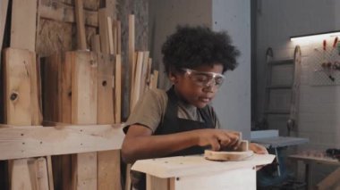 Afro-Amerikalı küçük çocuk marangozluk atölyesinde tek başına çalışırken kuş evinin yüzeyine çıkıyor.