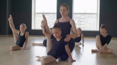 Genç koç, esnek bir kızın stüdyoda yerde otururken bacağını kaldırarak jimnastik yapmasına yardım ediyor.