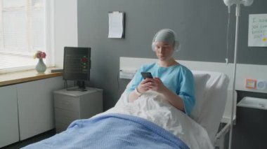 Kanserli genç bir kadın hastane koğuşunda telefon kullanırken ve müzik dinlerken kulaklıkla yatakta yatıyor.