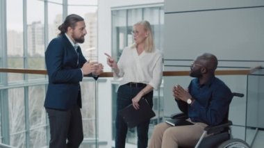 Çok ırklı işçiler, tekerlekli sandalyedeki adam ve ortakları modern ofis salonunda birbirleriyle konuşuyorlar.