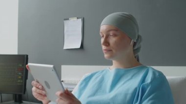 Kanser bölümünde yatakta yatarken dijital tablet kullanarak arkadaşlarıyla kulaklıkla iletişim kuran genç bir kadının orta boy fotoğrafı.
