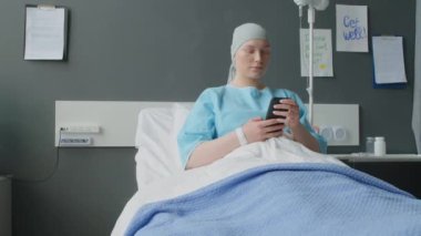 Orta ölçekli onkoloji bölümünden genç bir kadın hastanın modern hastanenin koğuşunda akıllı telefonuyla dinlenmesi.