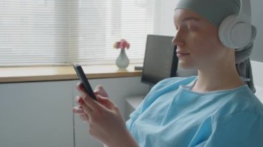 Kemoterapiden sonra kulaklıkla dinlenen genç bir kadın, koğuşta yatarken akıllı telefondan dinlemek için müzik seçiyor.