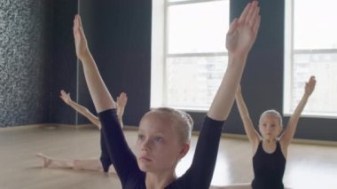 Küçük balerinler stüdyoda yerde otururken kollarıyla aşağı doğru dairesel hareketler yapıyorlar.