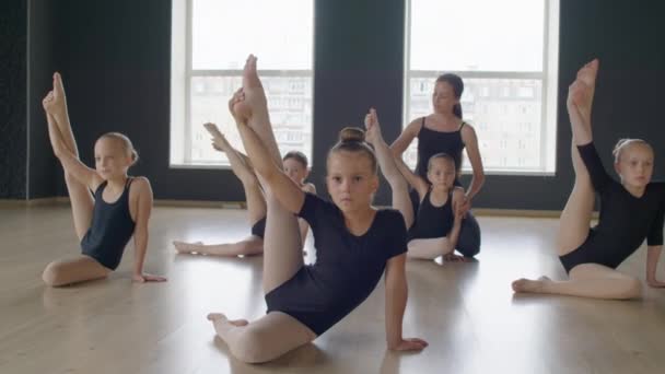 女性教師は 体操を行うこととリーチの女の子のチームを助け 床に座っている間に足を上げます — ストック動画