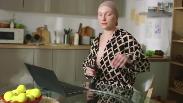 若いハゲの女性は ヘッドスカーフを離れて キッチンテーブルに座っているラップトップ上の友人のメッセージに答える — ストック動画