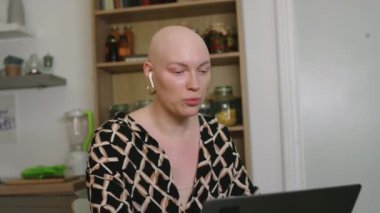 Kulaklıklı ve onkolojili neşeli kel bir kızın, mutfakta otururken bilgisayarından arkadaşını ararken orta boy fotoğrafı.
