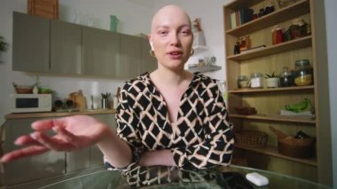 Onkolojisi olan kel bir kızın video görüşmesi yaparken ve mutfakta oturmuş kameraya bakarken görüntüsü.