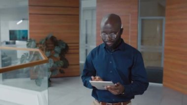 Modern ofis koridorunda yürürken dijital tablet kullanan ciddi bir Afrikalı Amerikalı ofis çalışanının orta boy fotoğrafı.