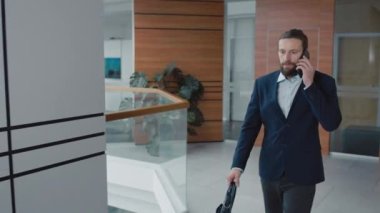 Beyaz takım elbiseli bir ofis çalışanı koridorda yürürken müşteriyle akıllı telefondan konuşuyor.