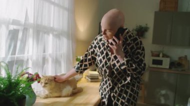 Orta boy, kel bir kadının evinde rahat bir şekilde akıllı telefonuyla konuşurken kızıl kedisini okşaması.