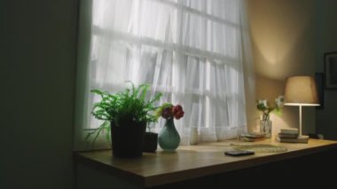İçi rahat oturma odasının içinde vazo, çiçek, bitki, kitap, akıllı telefon ve lamba dolu masa gündüz penceresinin yanında.