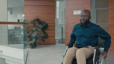 Orta uzunlukta, tekerlekli sandalyedeki neşeli Afrikalı Amerikalı adam modern ofis salonundaki iş arkadaşlarını selamlıyor.