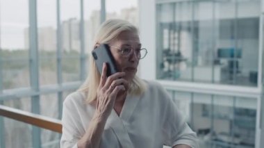 Yetişkin bir iş kadınının ofiste panoramik pencerelerle dikilirken müşterisiyle akıllı telefondan konuştuğu orta boy bir fotoğraf.