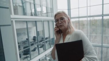 Modern ofis binasının asansöründe müşteriyle akıllı telefondan konuşan olgun bir iş kadınının orta boy fotoğrafı.