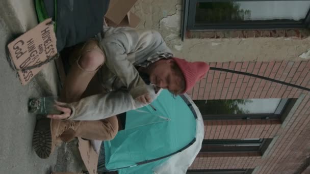 テントの隣に座って寄付を求めるタバコを抱えたホームレスの男の垂直ショット — ストック動画