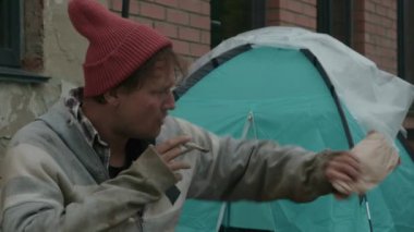 Orta halli bir sokak dilencisi sigara içmek için can atıyor. Barınağının ve dışarısının yanında otururken orta parmağını gösteriyor.