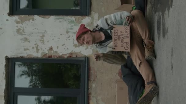 Вертикальний Знімок Безпритульного Чоловіка Знаком Просить Гроші Сидячи Землі Покинутому — стокове відео