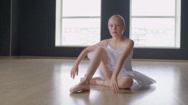 Pozitif küçük balerin portresi dans stüdyosunun zemininde bale kıyafetleriyle oturmuş panoramik pencere arka planında kameraya bakıyor.