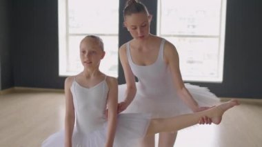 Stüdyoda bale dersinde esnetmesi için öğretmeninin yardımıyla bacaklarını bara koyan orta boy bir kız resmi.