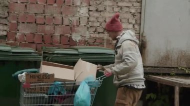 Alışveriş arabasıyla gündüz vakti çöp tenekelerini kazan evsiz adamın orta boy fotoğrafı.
