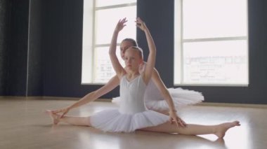 Beyaz elbiseli küçük bir balerin, ikiye bölünmüş bir şekilde oturuyor ve elleri yukarıda egzersiz yapıyor, öğretmen arkada oturuyor.