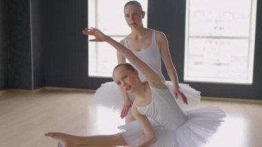 Orta uzunlukta beyaz etekli bir kız Barre 'de vücudunu ve kolunu çekiştirirken öğretmen ona yardım ederken.