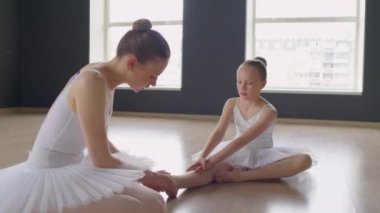 Genç balerin, bale dersinde yerde otururken kızların bileklerini sıkarken orta boy bir poz.