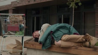 Ofis binasının yanındaki bankta uyuyan evsiz bir erkeğin orta boy fotoğrafı.