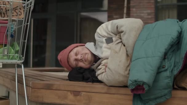 中等比例的可怜的流浪汉 穿着不整洁的衣服睡在购物车旁边的长椅上 他的东西都在房子附近 — 图库视频影像