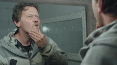 Evsiz bir erkeğin umumi tuvalette aynanın önünde yüz temizliği yaptığı orta boy bir fotoğraf.