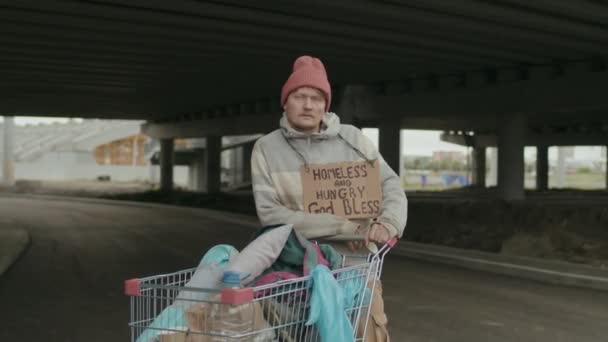 带有胸前标志的无家可归乞丐的画像 他们站在桥下 用购物车看着镜头 — 图库视频影像