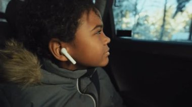 Dışarıda karlı manzarası olan bir yere arabayla giderken Afrika kökenli Amerikalı bir çocuğun kablosuz kulaklıkla müzik dinlemesi.