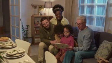 Çok nesilli kültürler arası aile, oturma odasındaki şenlik masasının yanındaki kanepede otururken dijital tablette videolar izliyor.