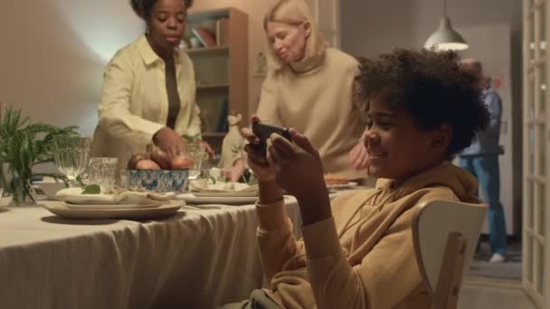 アフリカ系アメリカ人のティーンエイジャーがお祝いのディナーにテーブルを提供しながらスマートフォンで遊ぶことに焦点を当てる — ストック動画