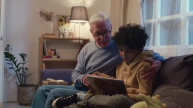 Orta boy, gözlüklü beyaz bir büyükbaba ve Afro-Amerikalı bir torunun birlikte kanepede otururken dijital tabletten bir şeyler izlediği bir fotoğraf.