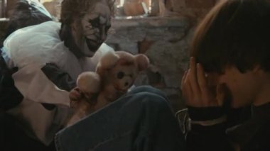 Orta ölçekli palyaço manyağı, kaçırılan kızı oyuncak ayıyla yerde otururken korkutur.