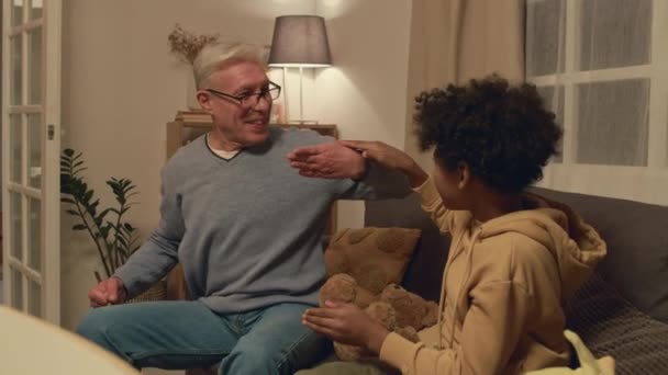 リビングルームのソファに座っている手の消えた指で焦点を当てたコーカサスのおじいちゃんを見ているアフリカ系アメリカ人の孫の中型ショット — ストック動画
