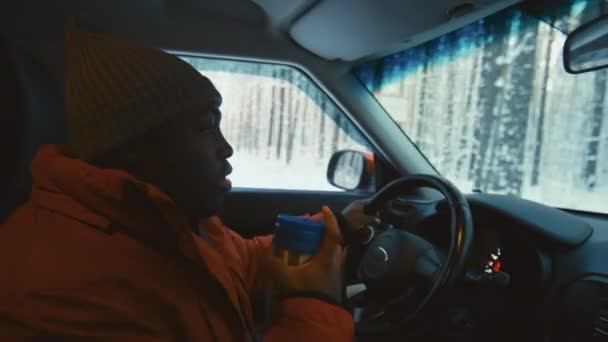 穿着保暖衣服和帽子开车的黑人男子的侧视图 在旅途中与坐在后座上的人交流 喝热饮 — 图库视频影像