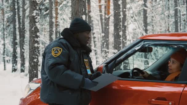 雪に覆われた森で車の女性と話す手のフォルダと暖かい制服で男性のアフリカ系アメリカ人の交通警官の中型ショット — ストック動画