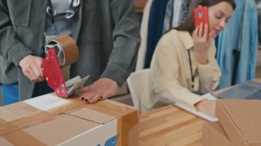 Başka bir işçi işyerindeki akıllı telefondan müşterileri ararken çevrimiçi mağaza müşterilerine gönderilen kutuları paketlemek için seçici bir odak noktası