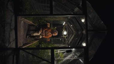 Sandalyeye oturup çay içen ve serada domates yapraklarına bakan çiftçinin dikey görüntüsü.