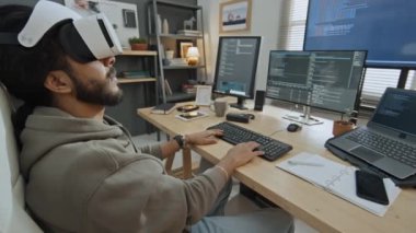 Sweatshirt 'lü genç bir erkek yazılım geliştiricisinin orta boy fotoğrafı, ev ofisinde sakallı, boyutsal model için bilgisayar kodu yazıyor, ve VR gözlükleri ile sonuçlara bakıyor.