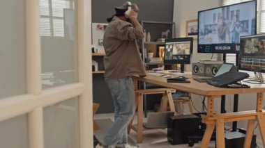 Kulaklıklı Ortadoğulu erkek tasarımcının evdeki iş istasyonuna doğru yürüdüğü ve profesyonel kurgu yazılımında video kliplerini işlemeye devam ettiği, müzik dinlerken tam kare.