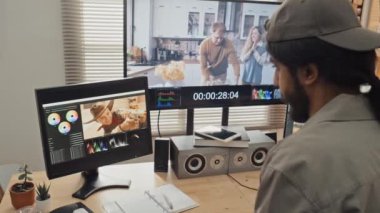 Evde masa başında duran serbest çalışan erkek tasarımcının orta boy arka plan görüntüsü ve profesyonel düzenleme yazılımında geniş ekranlı bilgisayardaki ham görüntü kliplerindeki renkleri düzeltme